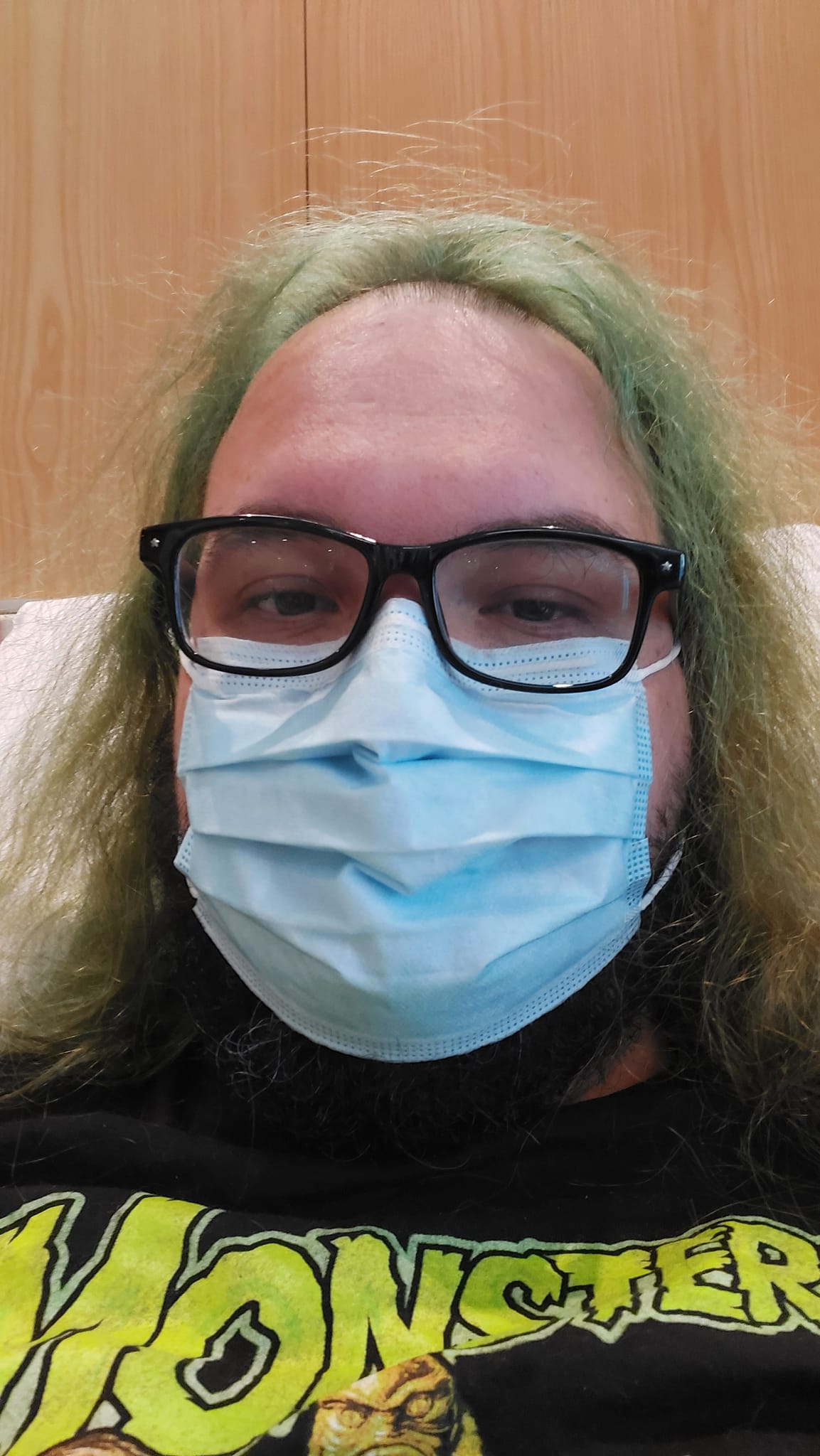 A man with green hair and a beard sits in a nondescript room with a surgical mask